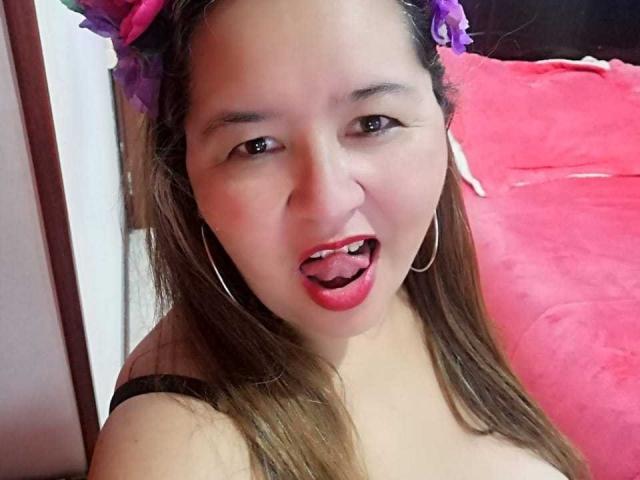 AmbarSimmons has light brown skin, long brown hair, purple flower headband, hoop earrings, red lipstick, tongue out, black strap top, and visible tits in a selfie.