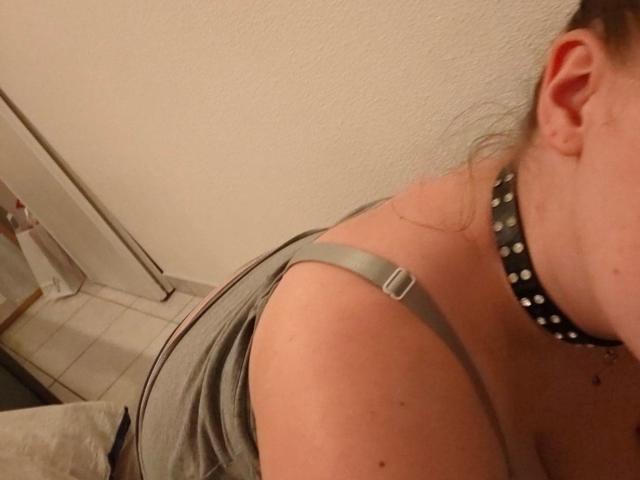 Le haut du corps de FrancaiseLynne, montrant une bretelle grise, un collier en cuir noir avec des clous métalliques, et son oreille, dans une pièce faiblement éclairée. Sa tête est légèrement tournée vers la droite..