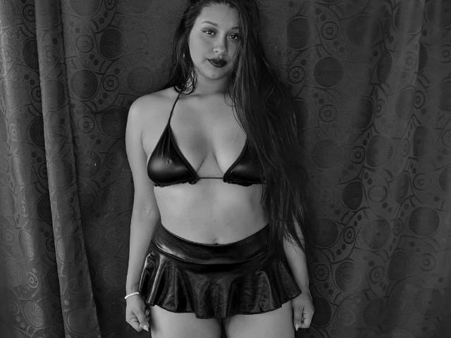 SharolXD with long wet hair, wearing a black shiny bikini top and a matching mini skirt, standing against a dark patterned curtain. Her tits are small, her ass is cute.