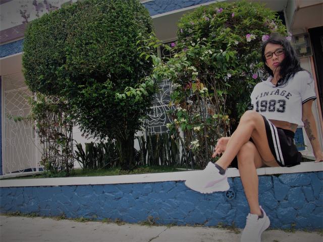 Daslyroze69 con gafas, cabello negro, top corto blanco con la inscripción "1983", pantalones cortos negros, zapatillas blancas, sentada en un muro de piedra azul, con las piernas cruzadas, un arbusto verde y frondoso detrás..