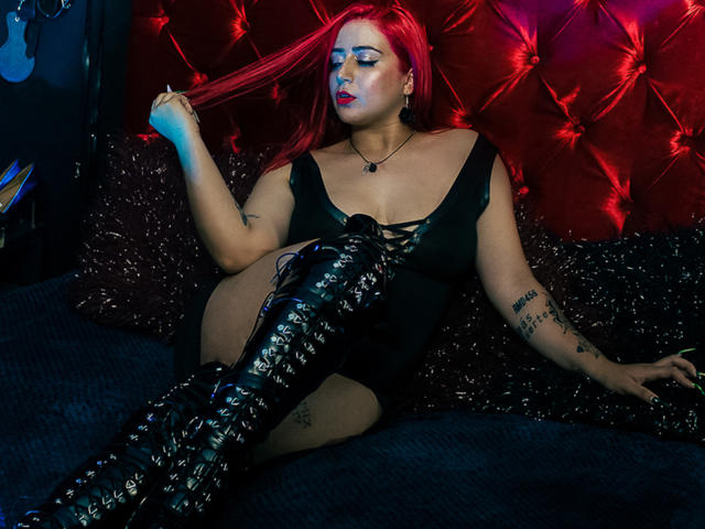 AprilDoel with red hair, black lace-up boots, and black dress, holding her hair, sitting on a dark bed, tattoos on arms, red tufted wall in the background.
