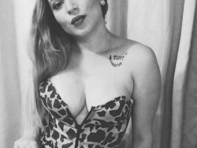OliviaMara mit langen Haaren, die ein trägerloses Kleid mit Leopardenmuster trägt, wobei ihre großen Brüste sichtbar sind, ein Tattoo an der rechten Schulter, schwarzer Lippenstift und Ohrringe, auf einem Schwarz-Weiß-Foto..
