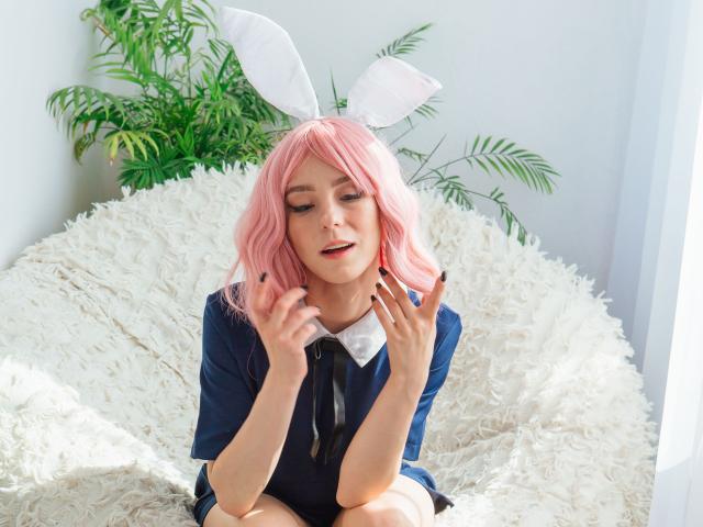 MissAstravert with pink wig, white bunny ears, dark blue dress, and black nail polish, sitting on white fluffy chair, hands near face, slightly open mouth, looking down.