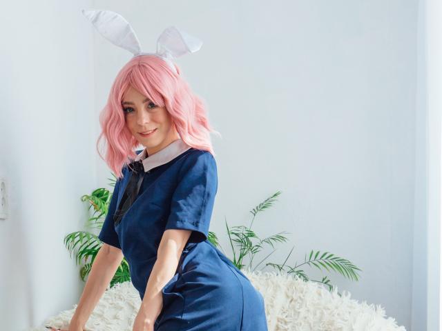 MissAstravert com cabelo rosa ondulado, orelhas de coelho brancas, vestido azul, gravata preta, gola branca, apoiada em uma superfície branca e fofa, plantas verdes ao fundo, sorrindo para a câmera, fofa..