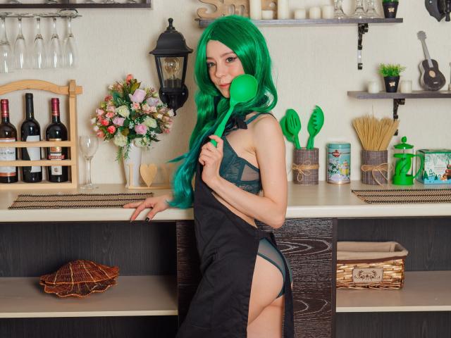 MissAstravert con parrucca verde, che tiene un cucchiaio verde, indossa un abito nero con spacco laterale, mostrando biancheria intima verde e glutei, in cucina con bottiglie di vino, fiori e utensili..