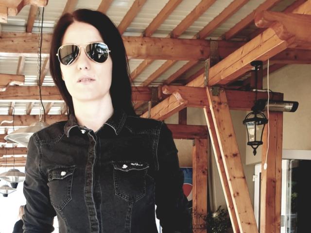 JoliDanielle with black denim shirt, buttoned, and reflective aviator sunglasses, has pale skin and straight black hair, standing under a wooden roof with a lantern hanging.