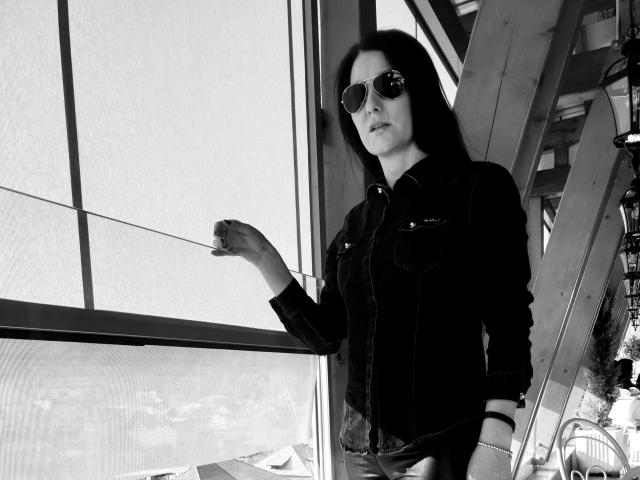 JoliDanielle with dark hair, wearing sunglasses and a black denim jacket, stands near a window, right hand on the glass, looking slightly to the left. Black-and-white photo.