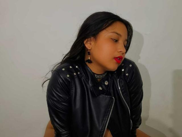 KailySeinsHot with long black hair, red lipstick, black leather jacket, and stud earrings, sitting, looking to the side, showing her ass slightly.