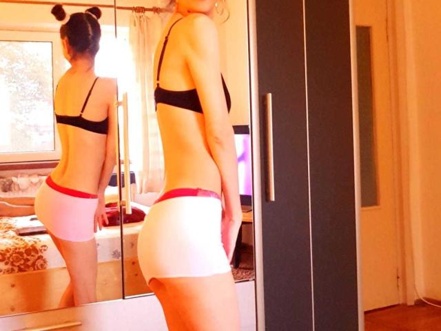 MelaniaTom with black bra, white and pink butt shorts, standing in front of mirror showing her back and ass. Her hair is in two buns. Bedroom.