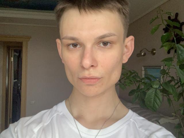 SmokerGuy hat ein blasses Gesicht, kurze braune Haare, braune Augen und trägt ein weißes Hemd mit einer silbernen Halskette. Sein Gesichtsausdruck ist neutral, er blickt direkt in die Kamera..
