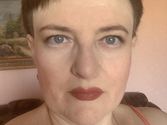 MargieEvans has short brown hair, blue eyes, and red lipstick. She's looking directly at the camera with a slightly serious expression. Her pale skin and small nose are visible.