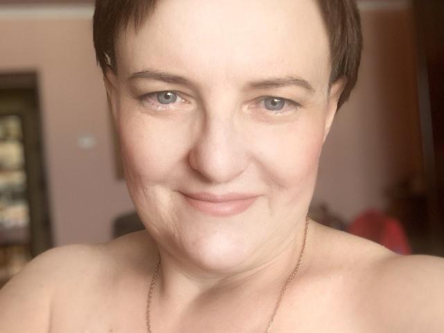 MargieEvans has short brown hair, light skin, blue eyes, and a subtle smile. She's shirtless, wearing a thin gold necklace, with visible small tits and a slightly chubby face.