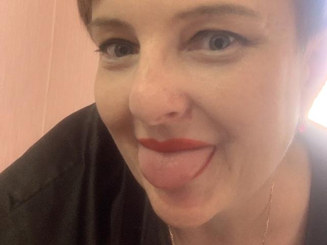 MargieEvans has short brown hair, blue eyes, and red lipstick. She's sticking her tongue out with a playful expression, wearing a black shirt and a gold necklace.