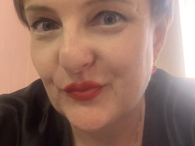 MargieEvans has short brown hair, blue eyes, and red lipstick. She's wearing a black top with a pink earring and a gold necklace. Her face is slightly puffy.