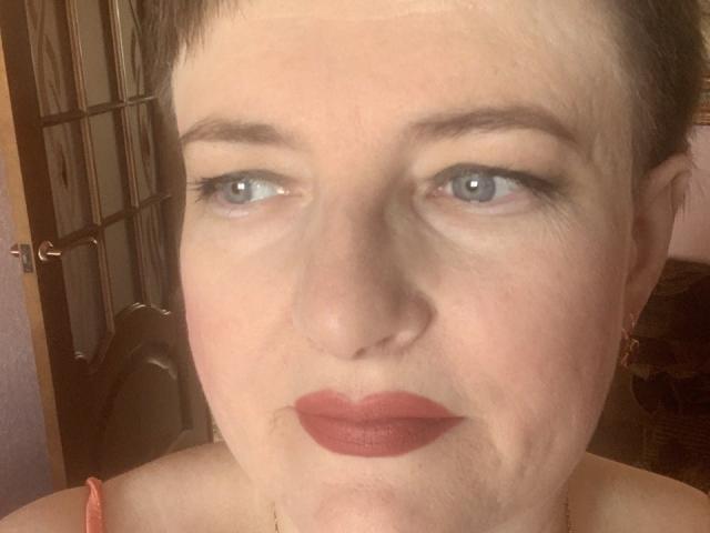 MargieEvans has pale skin, blue eyes, short brown hair, red lipstick, and small earrings. Her face shows slight wrinkles and a neutral expression. She's wearing a pink strap.