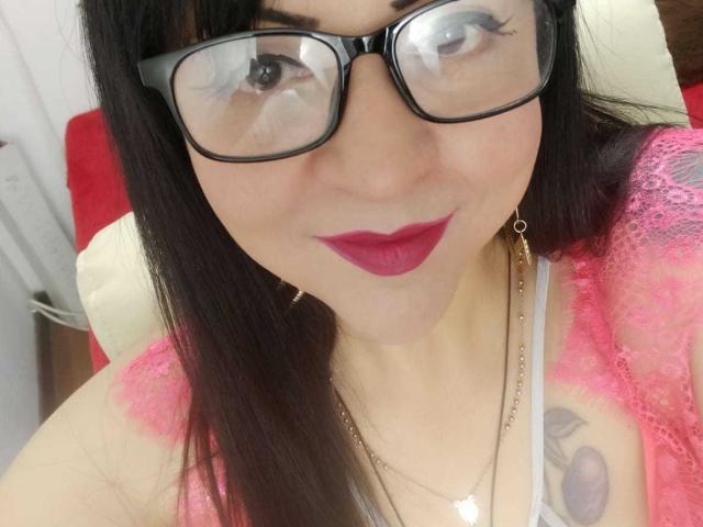AlishX with glasses, dark hair, pink lipstick, and red eyeshadow, wearing a pink lace top, showing a tattoo on her chest, smiling at the camera.