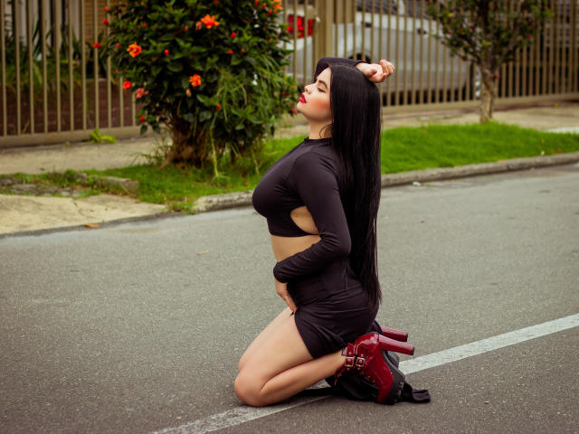 BrithneyLee kneels on a street, wearing a black crop top and mini dress, with red high heels, her hand on her ass, long black hair, eyes closed, lips red.