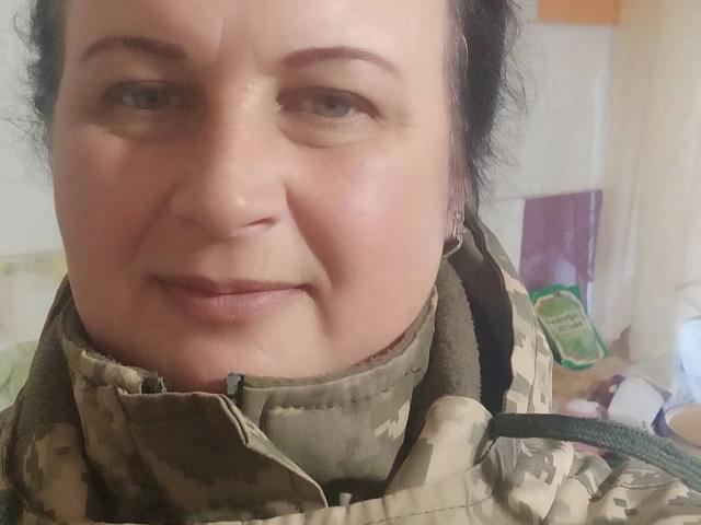 GinaMarlyn with a slightly smug expression, wearing a camouflage jacket, has pale skin, green eyes, and dark brown curly hair, showing a small nose and subtle smile.