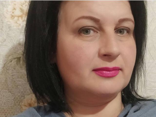 GinaMarlyn tem pele clara, cabelo preto na altura dos ombros, olhos verdes e batom rosa. Ela está vestindo uma blusa cinza, olhando ligeiramente para o lado com um sorriso discreto..