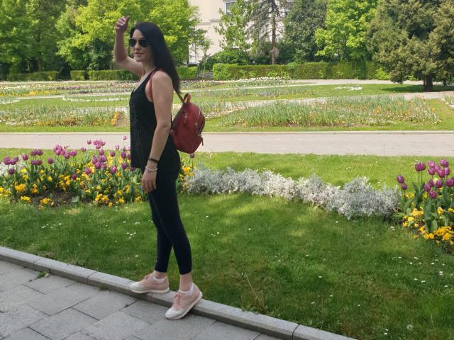 JoliDanielle with black hair, sunglasses, black tank top, black leggings, pink sneakers, red backpack, standing on sidewalk, waving, green garden with flowers and grass in background.