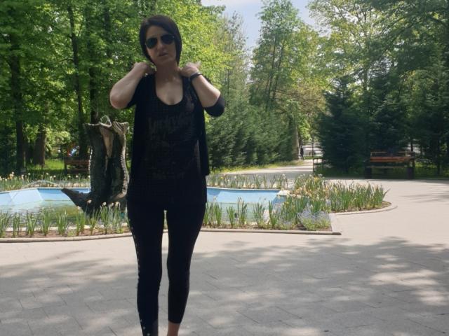 JoliDanielle stands in a park, wearing black lace top, black pants, and sunglasses, with hands on shoulders, showing her small tits and black wristband, in daylight.