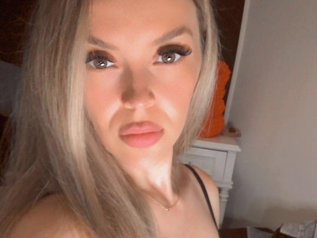 SophieDeee has blonde hair, brown eyes with long lashes, and pink lips. She's wearing a black top, and her tits are slightly visible. Her face is centered in the photo.