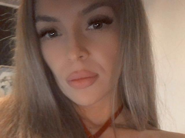 SophieDeee has long, straight brown hair, dark eyebrows, and full pink lips. She wears a red necklace, with subtle makeup highlighting her eyes. Soft, warm lighting.