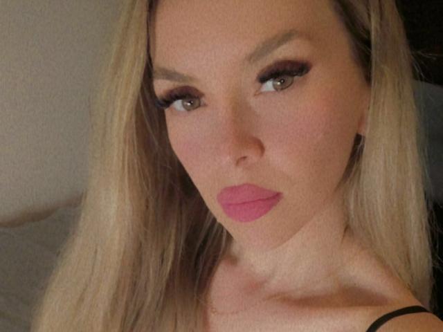 SophieDeee with blonde hair, brown eyes, and pink lipstick, showing off her tits with a slight pout. Her makeup includes long eyelashes. She's wearing a black strap.