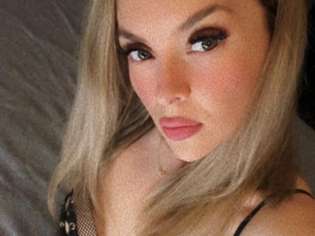 SophieDeee has blonde hair, brown eyes, and pink lips, wearing a black lace top with fishnet details, looking up with a slight pout. Her tits are partially visible.
