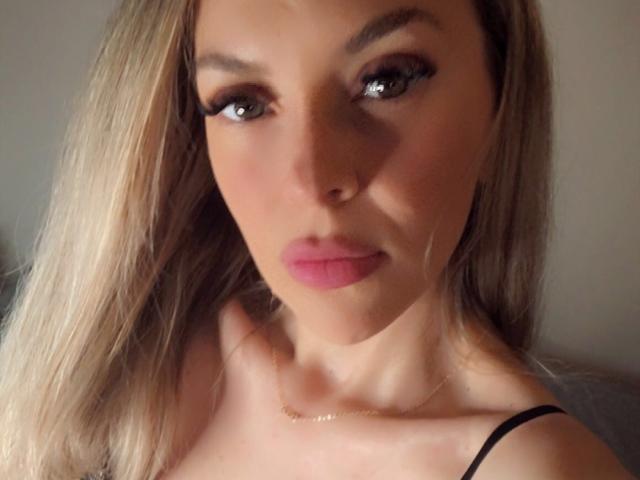 SophieDeee has blonde hair, green eyes, and pink lipstick. She's looking at the camera with a slight pout, wearing a black strap, and a thin gold necklace.