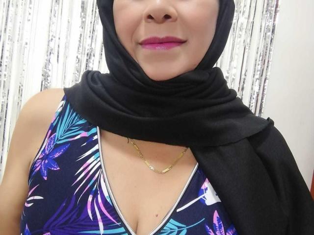 Acadia tiene senos de tamaño medio, lleva un hijab negro, una blusa floral morada y azul, un collar dorado y lápiz labial rosa, con un pañuelo negro sobre el hombro, y está sonriendo..