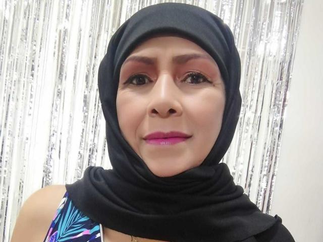 Acadia, com um hijab preto, sombra rosa, batom roxo e uma blusa estampada em azul, sorrindo para a câmera. Suas nádegas não estão visíveis. Ela está usando um lenço preto..