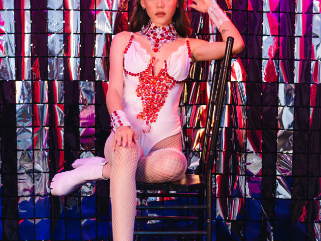 Jazmin Wells, avec une poitrine ornée de bijoux rouges, un body en dentelle blanche, des bas en résille, des bottes blanches, assise sur une chaise, tenant le dossier de la chaise, fond argenté et rouge, motif de grille sombre..