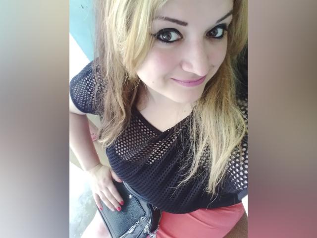 KamiaudX mit blonden Haaren, einem schwarzen Netzoberteil, rosafarbenen Shorts und roten Nägeln, lächelt in die Kamera, hält eine schwarze Tasche und steht in einem leicht unscharfen Bild..