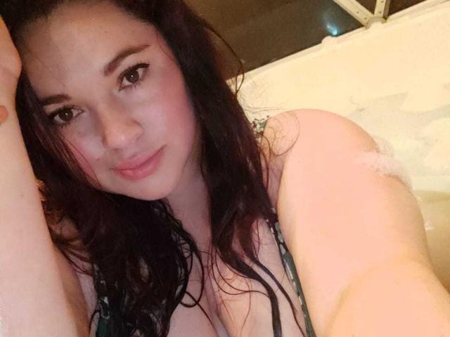 LissaClaire, com o cabelo escuro e molhado, bochechas levemente rosadas e um sorriso discreto, aparece em uma banheira, exibindo seus seios. Ela está vestindo um top de biquíni preto com estampa..
