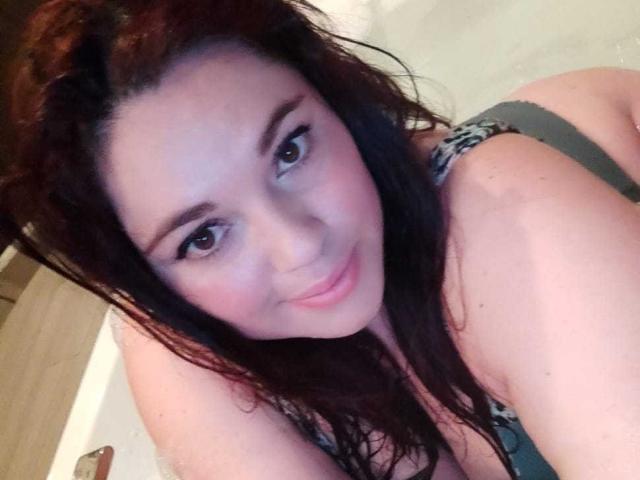 LissaClaire, com o cabelo castanho molhado, sorrindo, usando um sutiã verde, mostrando seus seios e nádegas em uma selfie. Seus olhos castanhos estão olhando para a câmera..