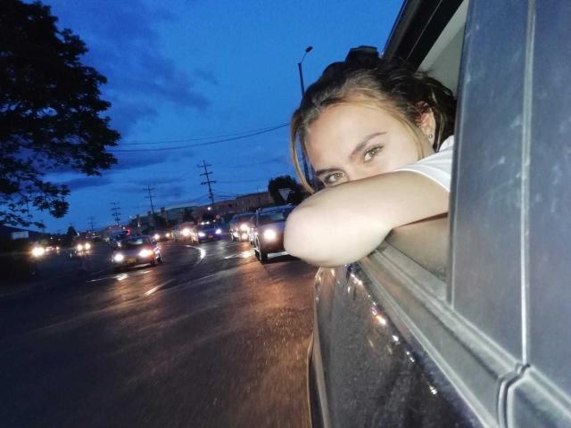 Le visage de MolySmith apparaît à la fenêtre d'une voiture, regardant sur le côté avec ses yeux verts, ses cheveux bruns attachés en queue de cheval, et portant une chemise blanche, lors d'une soirée pluvieuse..