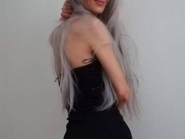 MelaniaTom with long, silver-gray hair, wearing a black, strapless dress, showing her slim back and small ass, smiling with one arm raised, in a simple, light-gray background.