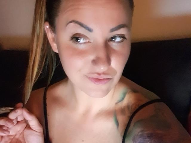 Una ragazza con un'aria sensuale, capelli castano chiaro raccolti in una coda di cavallo, che si tiene i capelli, ha tatuaggi sulla spalla destra e indossa una maglietta nera, sorridendo con le labbra leggermente arricciate..