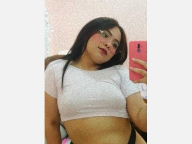 Samantha Velez, con cabello negro largo, usando gafas, una camiseta blanca corta y sosteniendo un teléfono rosa, mostrando sus pechos y una pequeña parte de su trasero en un selfie frente al espejo..