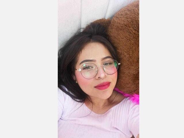 Samantha Velez, mit runder Brille, rosafarbenem Lippenstift und einem süßen Lächeln, liegt mit einem braunen Stofftier da und trägt ein rosa-weißes Hemd..