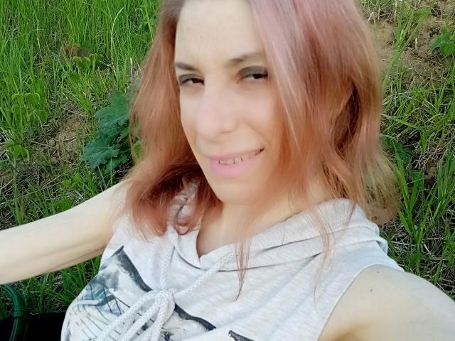 A selfie of ASexyLady with pinkish hair, smiling with slightly visible teeth, wearing a gray sleeveless top, standing in green grass. Her tits are small.