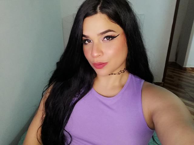AntonelaPark hat langes, schwarzes Haar, ein violettes Top ohne Ärmel, eine Halskette und pinken Lippenstift mit Eyeliner im Winged-Stil. Ihre kleinen Brüste sind sichtbar. Sie macht ein leicht schmollendes Gesicht auf einem Selfie..