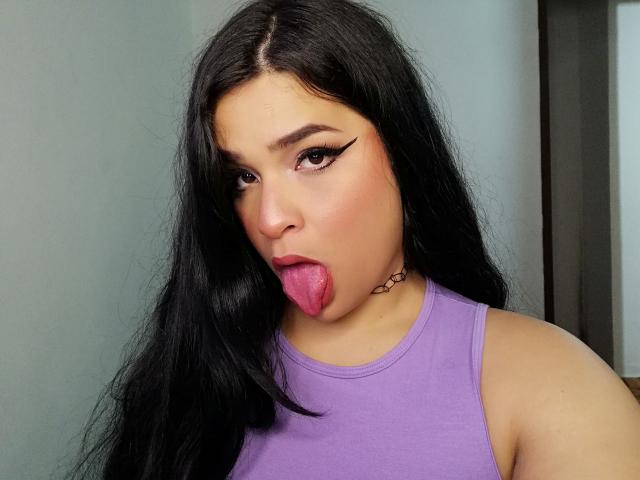 Antonela, con lunghi capelli neri, una canottiera viola e una collana, che mostra la lingua rosa, con un trucco audace sugli occhi (eyeliner ad ali di farfalla) e che guarda la fotocamera..