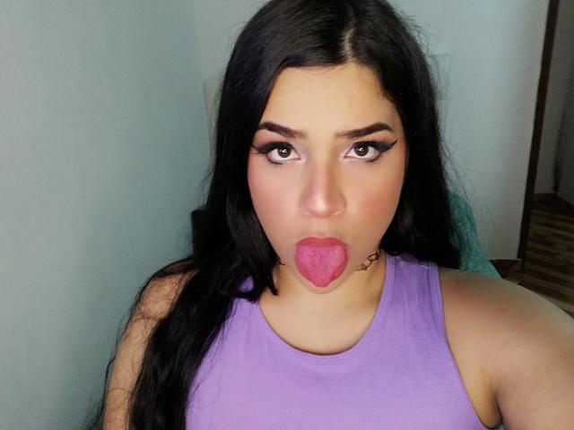 Antonela, con cabello negro largo, una camiseta morada y la lengua afuera, mostrando un tatuaje rosa en forma de corazón debajo de su boca. Tiene cejas pobladas y delineador de ojos alado..