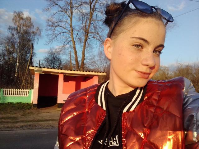 Solnushko69 mit heller Haut, braunen Haaren zu Dutt gebunden, trägt blaue Sonnenbrillen auf dem Kopf, eine rote, glänzende, gesteppte Jacke und ein schwarz-weiß gestreiftes Hemd. Sie steht vor einem rosa und grünen Gebäude..
