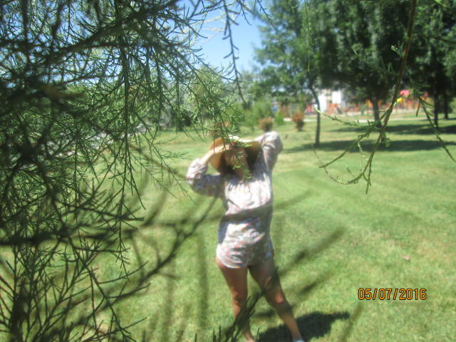 FacialEjac, with arms raised, stands in grass, wearing a white, patterned dress, partially obscured by green tree branches. Date stamp in bottom right corner.