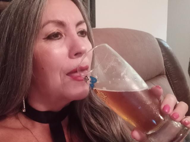 Eva Rodinson, con lunghi capelli castani, un collare nero, unghie rosa, che beve una birra alta, con le labbra intorno al bicchiere, con un leggero sorriso, tenendola con la mano destra..