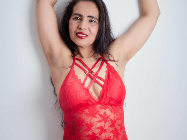MoxiaHot with arms raised, wearing red lace lingerie, showing small tits and armpit hair, smiling with red lipstick, long black hair, against a white wall.