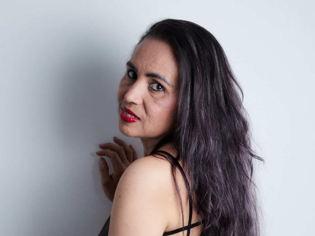 MoxiaHot has long, dark, wavy hair, red lipstick, and is looking over her shoulder with a slight smile, wearing a black, strappy top, showing her tits.